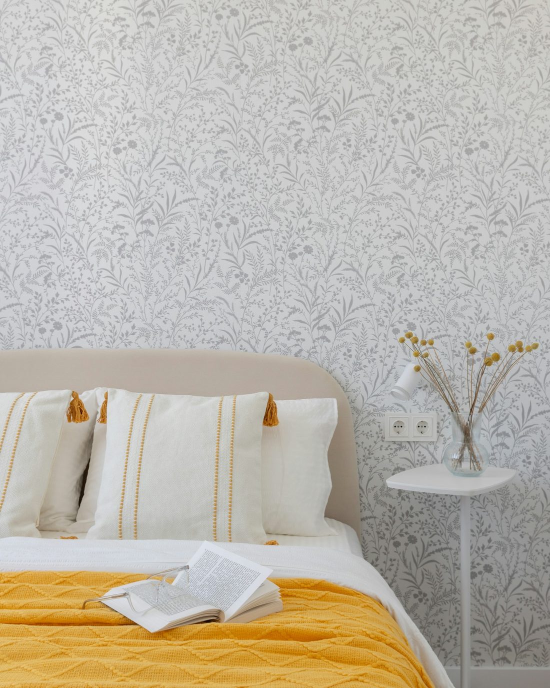 Calm bedroom with a pale grey botanical wallpaper behind a cream upholstered headboard, a mustard throw and a vase of dried flowers