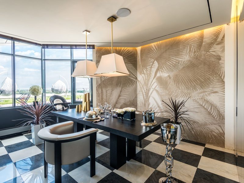 Dining room with a soft beige palm-frond mural lit by a cove light above a black table, brass pendants and a checkerboard marble floor