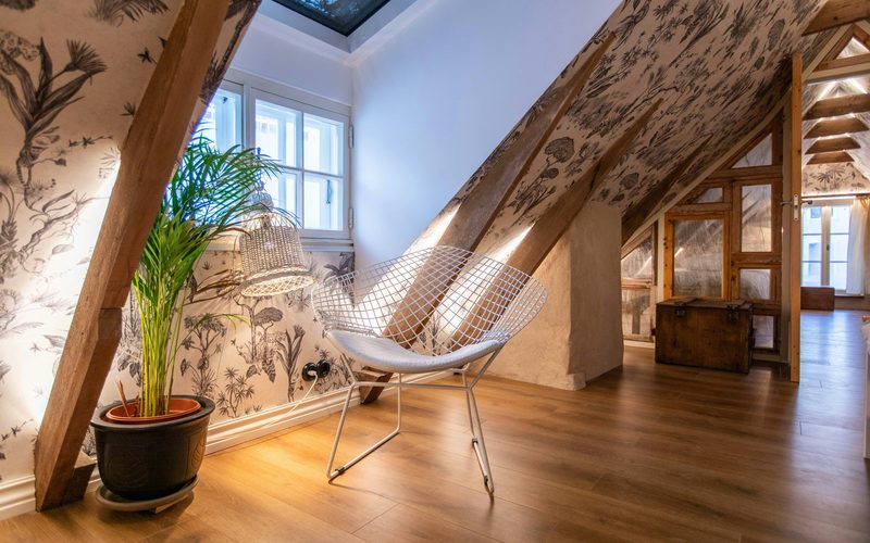 Attic room with black-and-cream jungle toile wallpaper on the sloped ceiling, exposed oak beams, a wire chair and a potted palm