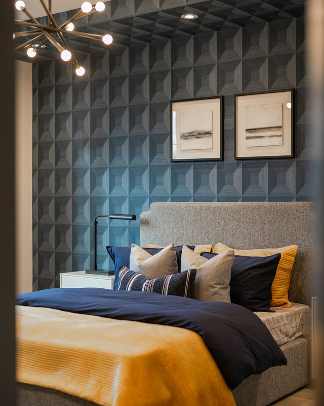 Bedroom with navy geometric pyramid-pattern wallpaper on the wall and ceiling, a grey bed with navy and mustard bedding and a brass chandelier