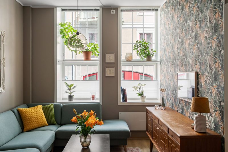 Living room with a sage and peach tropical-leaf accent wall beside taupe walls, a teal sofa and a mid-century sideboard
