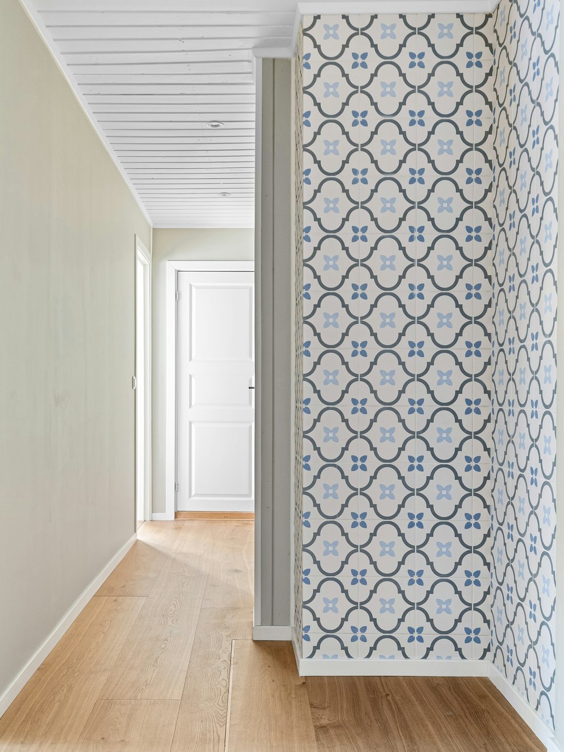 Bright hallway where a blue and white trellis-flower wallpaper wraps an outside corner beside a pale oak floor